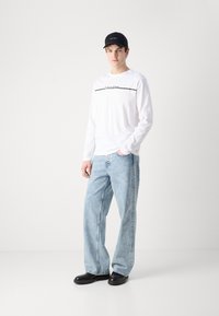 Weißes Langarmhemd mit schwarzem Logo und Liniendetails, hellblaue weite Denim-Jeans und schwarze Mütze, kombiniert mit schwarzen Schuhen.