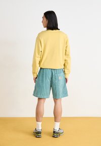Gelbes Sweatshirt, türkisfarbene Cordshorts mit Gesäßtasche, weiße Crew-Socken und graue Sneakers mit schwarzen und gelben Akzenten, von hinten betrachtet.