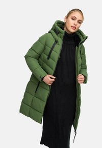 Green padded coat with a hood, zippered front, and black accents, worn over a ribbed black dress. Jacket features a quilted texture.