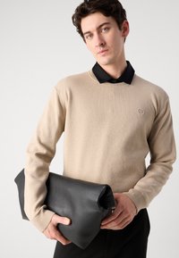 Beige kötött pulóver kerek nyakkivágással és logóval, alatta fekete galléros inggel párosítva. A modell egy fekete bőr táskával van.