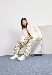 Nafúkaná krémová bunda v kombinácii so zelenými nohavicami s kvetinovým vzorom. Biele tenisky so svetlošedou podrážkou. Model sedí na kovovom objekte.