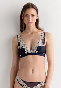 Soutien-gorge bleu marine avec un décolleté en V plongeant, bordure en dentelle beige et tissu en maille texturée. Se porte avec un slip assorti orné de dentelle.
