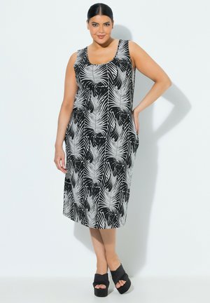 Kvinde iført en sort og hvid ærmeløs midikjole med abstrakte bladmønstre, stående med den ene hånd på hoften og sorte platformsandaler.