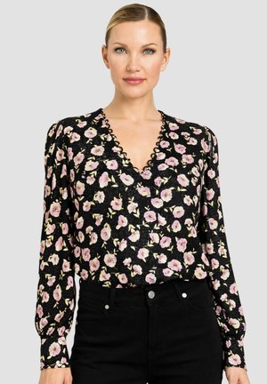 Donna che indossa una camicetta nera a maniche lunghe con stampa floreale rosa e scollo a V smerlato, abbinata a pantaloni neri a vita alta.