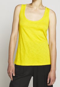 Henkilö, joka käyttää kirkkaan keltaista hihattomaa topia, jossa on pyöreä pääntie, ja löysiä mustia housuja, kuvattuna olkapäistä lantioon yksinkertaista taustaa vasten.