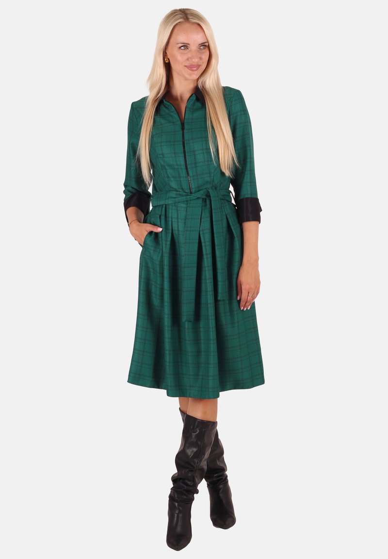 Grün karierter Kleid mit Reißverschluss vorne, langen Ärmeln und einem Bindegürtel. Verfügt über schwarze Akzente an den Bündchen und zwei seitlichen Taschen.