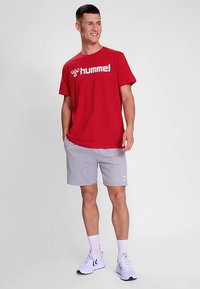 Rotes T-Shirt mit kurzen Ärmeln und weißem "hummel"-Logo, kombiniert mit grauen Shorts. Modell trägt weiße Socken und Sportschuhe. Schlichter Hintergrund.