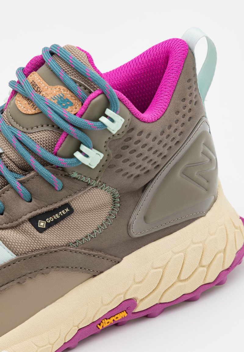 Zapatilla alta en marrón y verde azulado con acentos en rosa. Presenta el logo de Gore-Tex, parte superior texturizada y suela Vibram. Diseño ligero.
