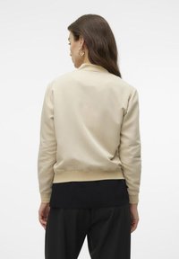 Beige Bomberjacke mit glatter Textur, geripptem Saum und Bündchen. Verfügt über eine schwarze gerippte untere Schicht, die über lockeren schwarzen Hosen getragen wird.