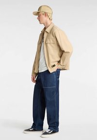 Beżowa kurtka, jasnoszara koszulka, ciemnoniebieskie luźne dżinsy i beżowa czapka. Buty sportowe z czarno-białymi akcentami. Widok z boku, jednolite tło.