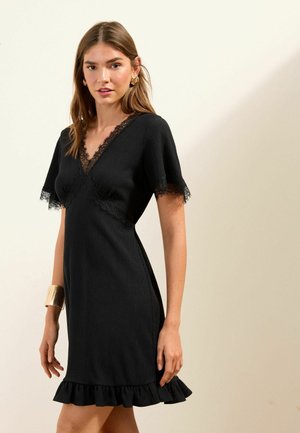 Frau in einem schwarzen, kurzärmligen Kleid mit Spitzenbesatz an Ärmeln und Ausschnitt, einem gerüschten Saum und einem goldenen Armreif am linken Handgelenk.