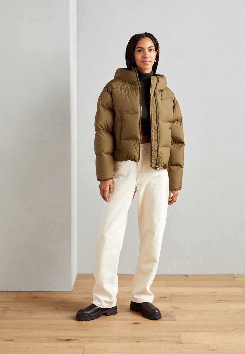 Olivgrüne Steppjacke mit einem cropped Design und Kapuze, kombiniert mit hellen, weiten Hosen und dunklen klobigen Stiefeln.