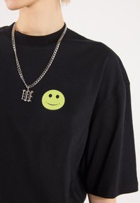 Juodas per didelis t-shirt su apvaliu žaliai citrinos spalvos šypsenėle ant krūtinės, derinamas su sidabrine grandinėlės apyranke su pakabuku.