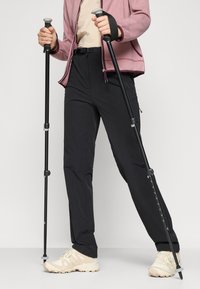 Čierne outdoorové nohavice so slim fit strihom, skombinované so béžovými turistickými topánkami. Model drží trekingové palice a má na sebe ružovú bundu na zips.