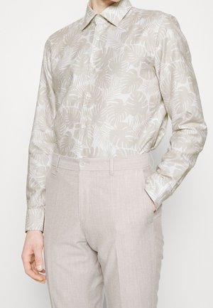 Homme portant une chemise beige à manches longues avec motif de feuilles, rentrée dans un pantalon beige clair sur mesure, une main dans la poche.