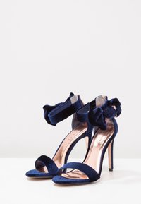 Marina sammet högklackade sandaler med öppen tå och spänne runt vristen. Har en rosett som accent och en metallisk roséguldig innersula.