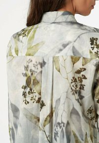 Lightweight, patterned blouse featuring a gray background with green and brown botanical prints, adorned with a classic collar and back pleat.