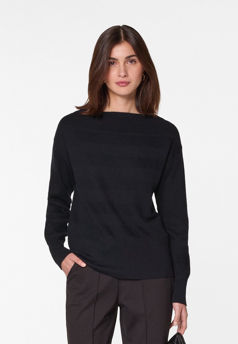 Pull noir en tricot avec un décolleté bateau, manches longues et de subtils détails de texture horizontale. Associé à un pantalon sombre.