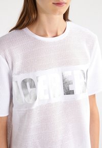 Bílý síťovaný tričko s širokým stříbrně metalickým potiskem s nápisem "ICEBERG" přes přední stranu. Krátké rukávy a kulatý výstřih.