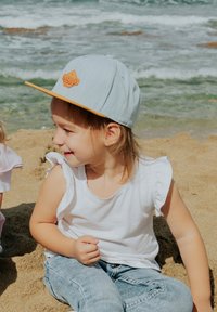 Light blue cap with a flat brim, orange logo patch, worn by a child in a white ruffled shirt and denim jeans sitting on sandy beach.