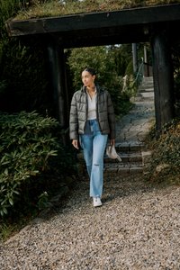 Szara pikowana kurtka, biała prążkowana bluzka, jasnoniebieskie dżinsy z dziurą. Idzie po kamiennej ścieżce otoczonej zielenią. Trzyma torbę.