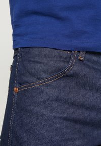Mörkblå denimjeans med femficksdesign, synlig orange sömnad och knappdetaljer. En blå bomulls-t-shirt är delvis synlig ovanför.