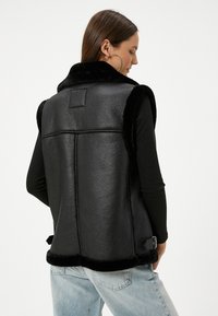 Schwarze Lederweste mit einem Kragen und Saum aus Kunstfell, die ein elegantes Design, einen nahtlosen Rücken und eine Schnallendetail an der Taille aufweist.