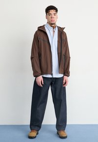 Brown lightweight jacket with a hood, zippered pockets, layered over a light blue striped shirt, paired with loose dark jeans and brown shoes.