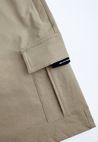 Beige stoffen shorts met een zijzak, gestikte details en een zwart label met "BLCK" in witte tekst. Gladde textuur, op maat gesneden pasvorm.