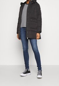 Osoba v černé prošívané bundě, černém a bílém pruhovaném tričku, modrých skinny džínách a černých vysokých teniskách, stojící na jednobarevném pozadí.