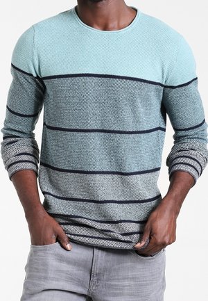 Mies, joka käyttää teksturoitua, pitkähihnaista villapaitaa, jossa on vaakasuuntaisia sinisen ja harmaan sävyisiä raitoja, yhdistettynä vaaleanharmaisiin housuihin.