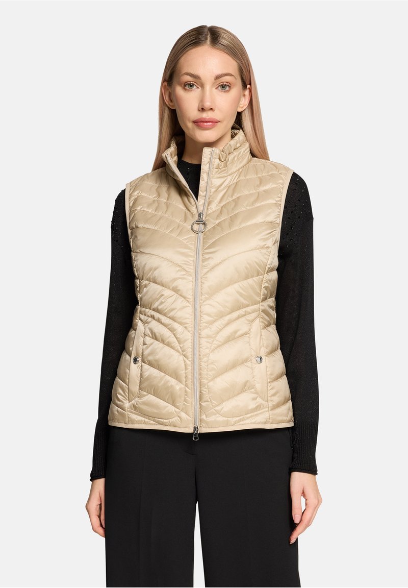 Veste matelassée beige avec col montant, fermeture éclair à l'avant et poches latérales. Portée par-dessus un pull noir avec une texture subtile.