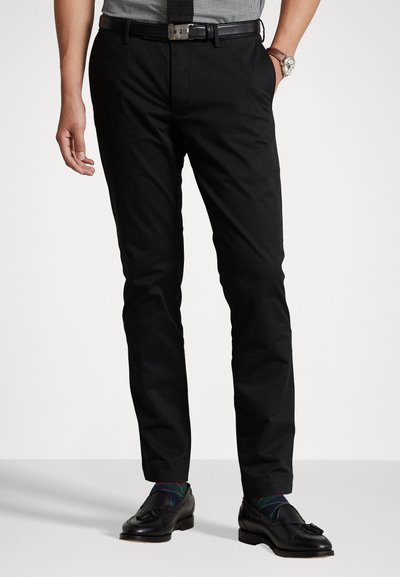 Pantaloni neri eleganti a vestibilità slim, realizzati in tessuto morbido; abbinati a scarpe nere lucide e calzini con fantasia. Presentano una cintura e un orologio.