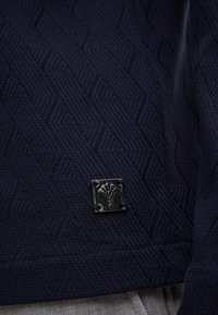 Pullover in maglia blu navy con un motivo a spina di pesce texturizzato e un piccolo emblema metallico con un simbolo medico sul lato inferiore.
