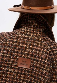 Gewebter brauner und burgunderfarbener Tweed-Mantel mit hohem Kragen, auf dessen Rückseite ein Lederschild "blue shadow" sichtbar ist. Ein brauner Filzhut ist ebenfalls zu sehen.