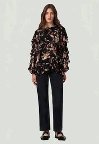 Blusa preta com flores, com mangas em camadas e babados, apresentando uma mistura de flores cor-de-rosa e creme. Combinada com jeans escuros de perna reta e sapatos pretos.