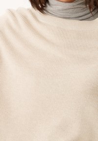 Beige gebreide trui met een geribd patroon en een wijde halslijn, over een lichtgrijze coltrui gedragen.