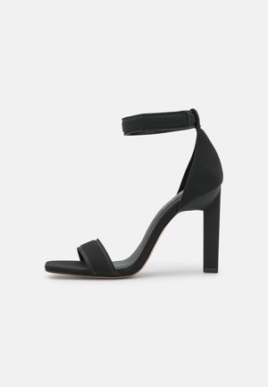 Schwarze High-Heel-Sandale mit Knöchelriemen, offenem Zehenbereich und Blockabsatz vor weißem Hintergrund.