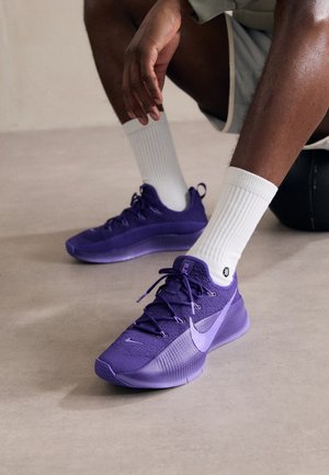 Personne portant des chaussures de course Nike violettes et des chaussettes blanches, assise sur un sol clair avec une main posée sur son genou.