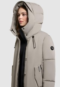 Beige pufjakke med en stor hætte, frontzip og trykknaplukninger, to sidelommer og branding på ærmet. Blødt, struktureret stof.