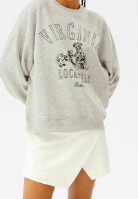 Šedá mikina s černou grafikou dvou psů a textem "VIRGINIA LOCATTEAM RICH." V kombinaci s bílou zavinovací sukní. Bavlněný materiál, volný střih.