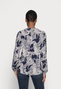Persona con capelli ricci corti che indossa una blusa blu navy con motivi paisley bianchi e jeans scuri, voltata di spalle su uno sfondo semplice.
