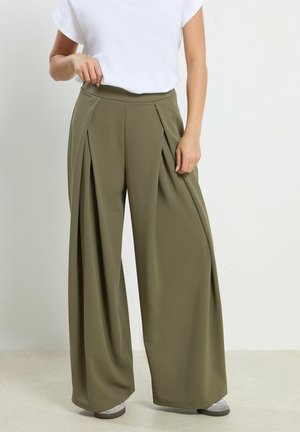 Donna con pantaloni plissettati a gamba larga verde oliva, sneakers bianche e top bianco a maniche corte, in piedi davanti a uno sfondo uniforme.