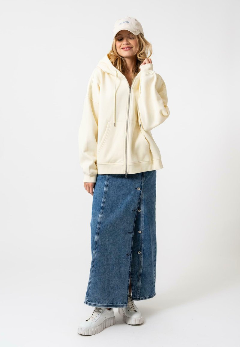 Sudadera amarilla clara con cremallera, falda larga de mezclilla azul hasta la rodilla con botones en el frente, zapatillas de plataforma blancas y gorra beige.