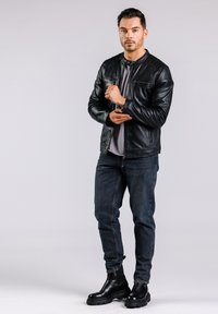 Chaqueta de cuero negra con detalle acanalado en los hombros, camiseta gris debajo, jeans oscursos y botas negras de suela gruesa. Modelo posando de pie.
