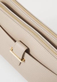 Bežová kožená taška s texturovaným povrchem, vybavená dvojitými zlatými zipy a zlatým akcentovým klipem. Kompaktní obdélníkový design.