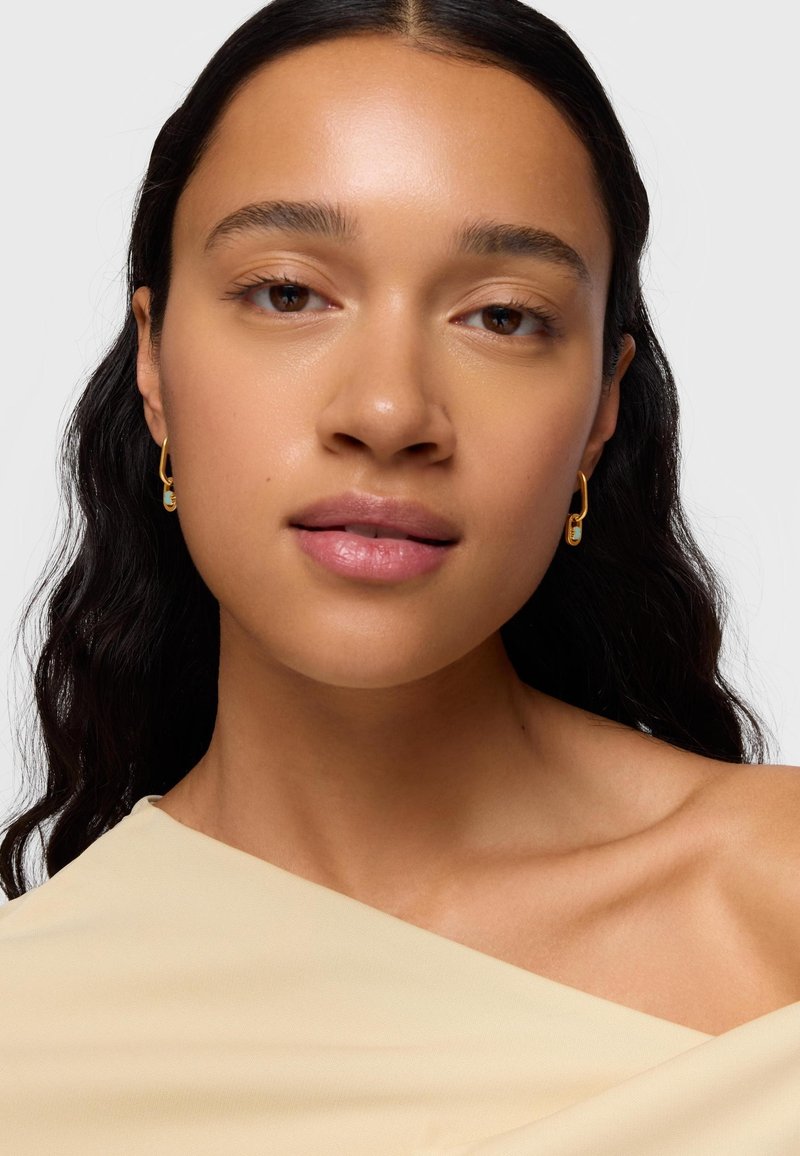 Femme à la peau lisse et aux cheveux ondulés foncés porte des boucles d'oreilles en or avec de petits détails turquoise et un haut beige décolleté épaules.