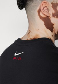 Svart sweatshirt med rund halsringning, med en vit Nike-logotyp och röd "AIR"-text på baksidan. Mjuk tygkvalitet, avslappnad design.