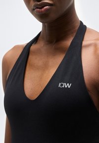 Κοντινό πλάνο ενός ατόμου που φοράει μαύρο τοπ με halter, βαθύ V λαιμόκοψη και λευκό λογότυπο "ICIW" στο στήθος.