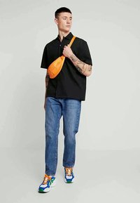 Camisa preta de manga curta, jeans azul claro com corte reto, bolsa transversal laranja brilhante, ténis multicoloridos com atacadores.